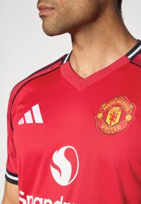 Červený dres Manchester United s výstrihom do V. Obsahuje biele logo Adidas, vyšívaný erb klubu a čierne pruhy na rukávoch.