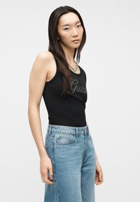 Jeune femme aux longs cheveux bruns portant un haut noir sans manches avec le logo "Guess" et un jean taille haute bleu clair, sur un fond uni.