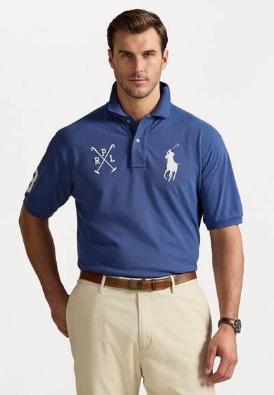 Blå polo med broderet hvidt logo og accenter, korte ærmer, en tre-knap lukning og et struktureret stof. Båret med beige bukser.
