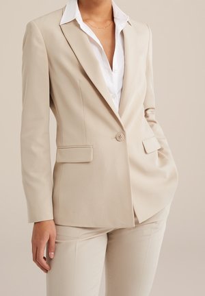 Femme portant un blazer beige sur mesure et un pantalon assorti, avec une chemise blanche à col, une main dans la poche, fond neutre.