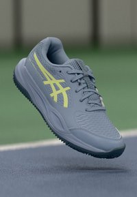 Graue Sportschuh mit einer strukturierten Oberfläche, gelben Akzenten und einem atmungsaktiven Mesh-Obermaterial. Verfügt über eine gepolsterte Sohle und ein Schnürdesign.