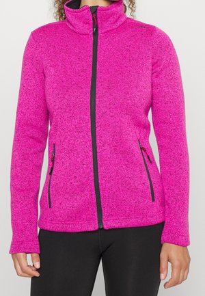 Frau trägt eine hellpinkfarbene Fleecejacke mit Reißverschluss und schwarzen Reißverschlüssen sowie schwarze Hose, steht vor einfachem hellem Hintergrund.