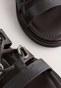 Sandales en cuir marron foncé avec larges brides et boucles métalliques argentées, montrées en gros plan sur un fond clair.