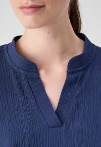 Marineblå bluse med en teksturert overflate, med ståkrage og en subtil V-hals. Myk stoff. Nærbilde av halsen.