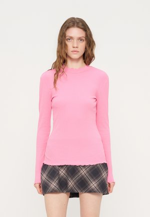 Ung kvinde med bølget hår iført en langærmet lyserød top og en sort ternet mininederdel, stående mod en ensfarvet hvid baggrund.