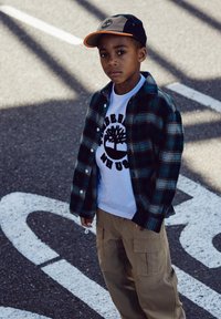 Un bambino indossa una camicia a quadri verde e blu sopra una maglietta bianca con un logo ad albero nero, abbinata a pantaloni cargo beige e un cappellino nero.