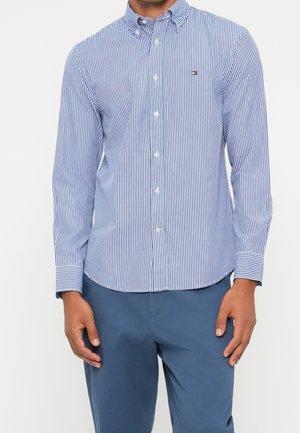 Mann trägt ein blau-weiß vertikal gestreiftes Hemd mit Knöpfen und blaue Hose, steht mit entspannten Händen an den Seiten.