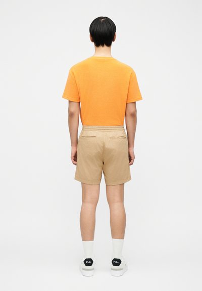 Άνδρας που στέκεται με την πλάτη, φοράει πορτοκαλί μπλουζάκι, μπεζ σορτς, λευκές κάλτσες και λευκά αθλητικά παπούτσια με μαύρα σημεία στη φτέρνα.