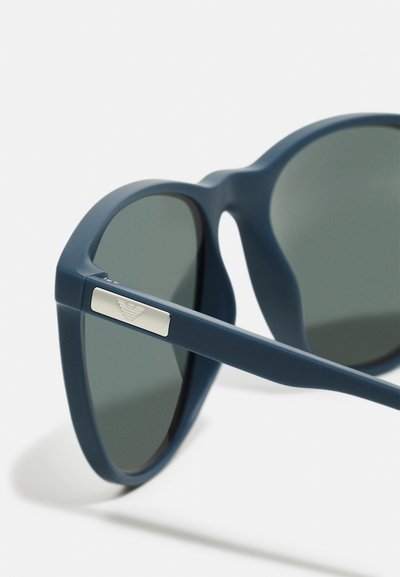 Occhiali da sole in plastica blu con lenti grigie. Presentano una superficie liscia e un accento argentato sulla stanghetta. Design angolare, leggermente sovradimensionato.