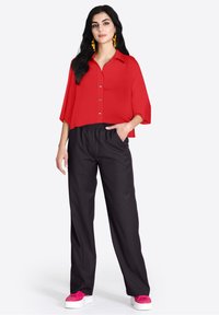 Blusa rossa a maniche corte con colletto abbinata a pantaloni neri a vestibilità comoda e sneakers rosa. La blusa presenta un design con bottoni.