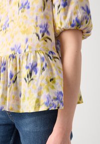 Gros plan d'une personne portant un chemisier jaune avec des motifs floraux bleus et des manches bouffantes, associé à un jean en denim bleu.