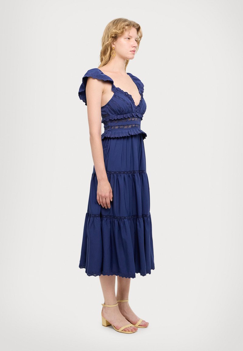 Navyblaues Kleid mit rüschierten Ärmeln, Spitzenakzenten, gestuftem Design und taillierter Taille. Tragen mit gelben Schnürsandalen.