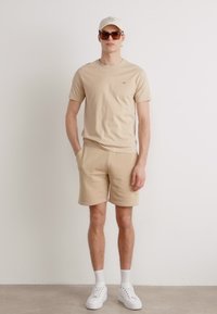 Beige T-shirt og shorts lavet af blødt stof. Tilbehøret inkluderer en lys kasket og solbriller. Sneakers fuldender det afslappede look.