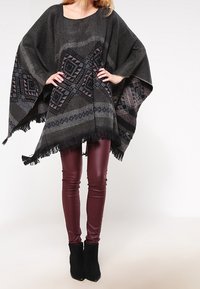 Černý a šedý tkaný pončo s geometrickými vzory, s třásněmi, nošené přes burgundské legíny a černé kotníkové boty.