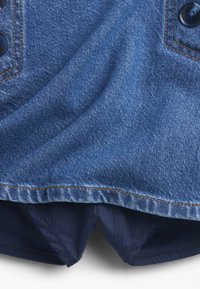 Sötétkék farmeranyag narancssárga varrással. A mintázat gombos részleteket és enyhén rojtos alsó szegélyt tartalmaz.