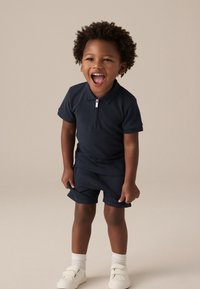 Polo marine avec col zippé, shorts assortis, et baskets blanches à fermeture velcro. Tissu doux et coupe décontractée adaptée aux enfants.