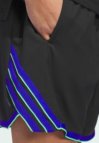 Pantalones cortos negros con acentos a rayas en púrpura y verde y un acabado de malla. Bolsillo lateral visible que muestra la textura de la tela transpirable.