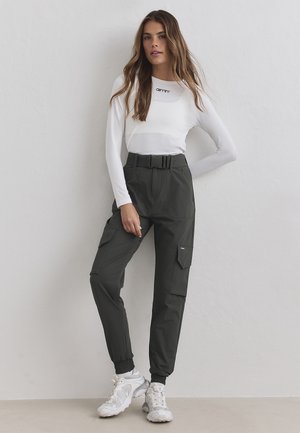 Femme appuyée contre un mur blanc, portant un haut blanc à manches longues, un pantalon cargo noir taille haute et des baskets blanches.