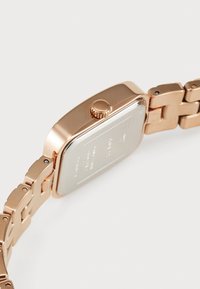 Ceas de aur roz cu carcasă rectangulară, coroana texturată și brățară metalică legată. Capac din sticlă plată și fundal gravat.