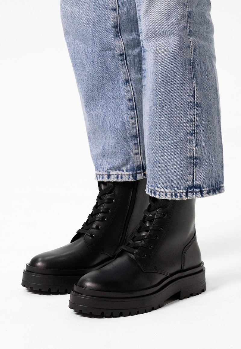 Bottes de combat en cuir noir avec une semelle épaisse, bout rond et lacets, assorties à un jean bleu clair retroussé.