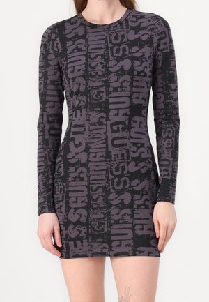 Kobieta nosząca dopasowaną, czarną sukienkę z długim rękawem, ozdobioną szarymi pionowymi tekstowymi wzorami. Sukienka ma długość powyżej kolan.