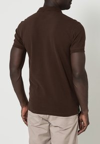 Polo marron avec une texture lisse, manches courtes et col. Détails de couture visibles et une petite étiquette à l'ourlet.