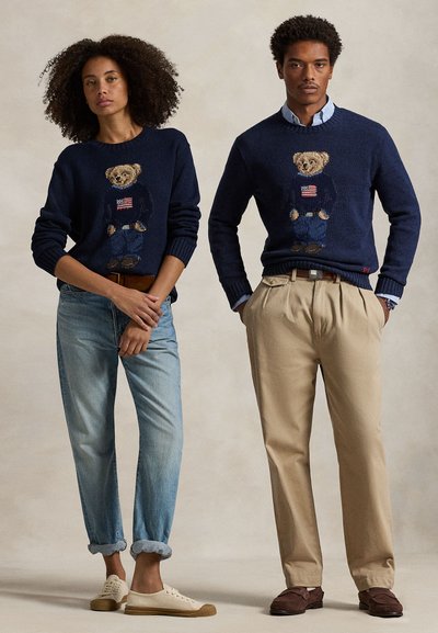 Woman and man standing, both wearing navy sweaters with teddy bear and American flag designs; woman in jeans and sneakers, man in beige pants and loafers.