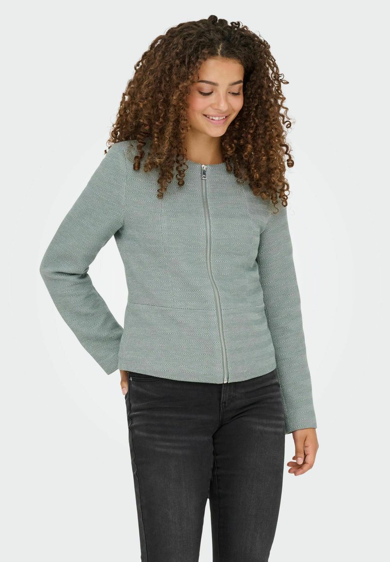 Ljust grön zip-up jacka med en strukturerad fiskbensmönster, långa ärmar och en figursydd design, bärs över mörka jeans.