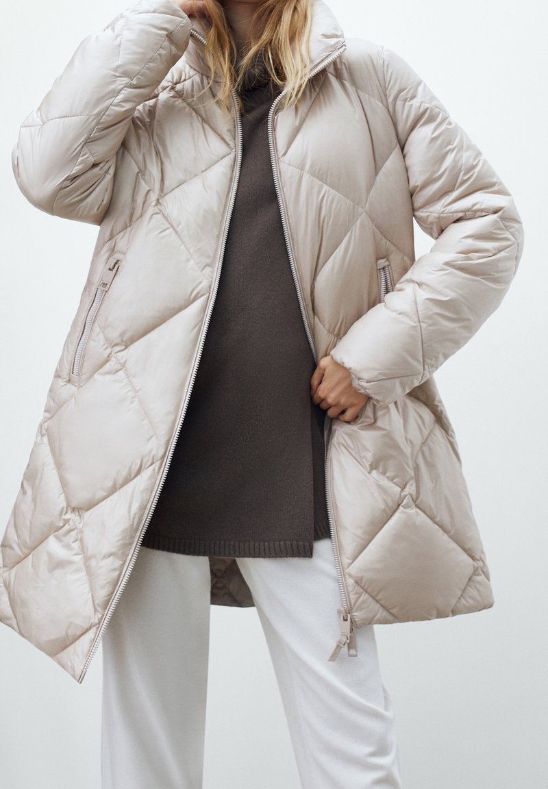 Personne portant un manteau matelassé beige sur un pull marron foncé et un pantalon blanc, en train d'ajuster la fermeture éclair du manteau.