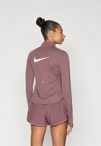 Μπλούζα μακρυμάνικη και σορτς Nike σε απόχρωση μωβ. Η μπλούζα διαθέτει ψηλό γιακά και λευκό λογότυπο στην πλάτη· τα σορτς έχουν λευκή διακόσμηση και λιτή λεπτομέρεια.