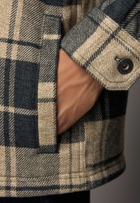 Kariertes Jackett in Beige- und Marineblau-Tönen, strukturierter Stoff und seitliche Tasche. Der Knopfverschluss am Ärmel verleiht zusätzliche Details.