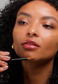 Femme aux cheveux noirs bouclés appliquant un fond de teint liquide sur le visage à l'aide d'un applicateur, avec des points de fond de teint visibles sur le front et les joues.