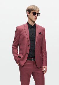 Piros-rózsaszín szabott öltöny fekete inggel és zsebkendővel. Slim fit fazon, kivágott hajtókák, és fekete napszemüveg jellemzi. Sima textúra.
