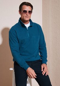 Teal Fleece-Pullover mit langem Arm und Viertelreißverschluss; kombiniert mit dunklen Hosen. Modell trägt Sonnenbrillen und sitzt auf einem Hocker.