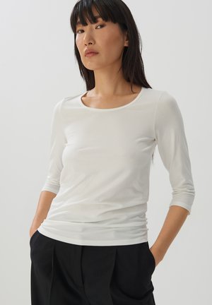 Chemise blanche ajustée à manches longues avec un décolleté en bateau et un tissu léger et doux. La chemise a des manches trois-quarts et un design sans couture.