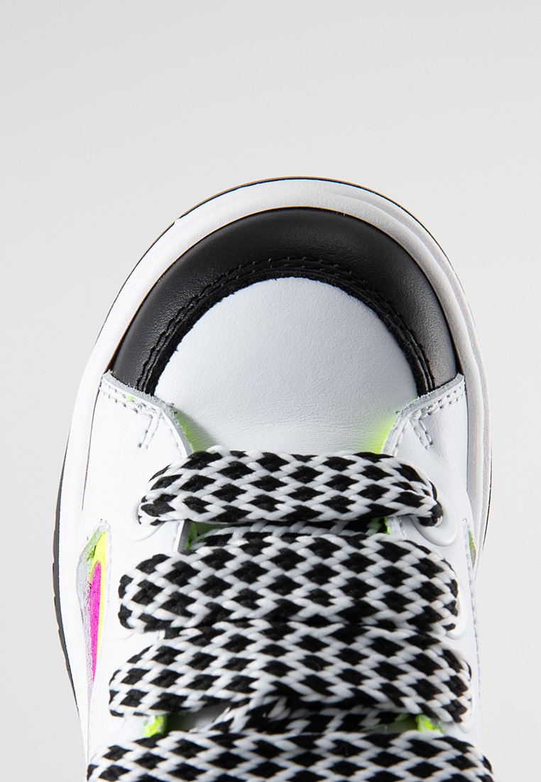Białe sneakersy z czarnymi akcentami, skórzany cholewka o strukturze, wyraźne czarno-białe sznurowadła w kratkę oraz mały neonowo-różowy detal na boku.