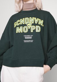 Felpa verde scuro realizzata in un tessuto morbido, caratterizzata da bold testo giallo e dettagli stampati. Ha una vestibilità rilassata e polsini a coste.