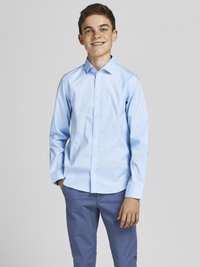 Chemise bleu clair à manches longues avec un col boutonné ; tissu uni et coupe classique. Associée à un pantalon bleu, les mains dans les poches.