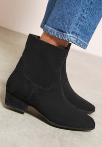 Melnas zamšādas potītes zābaki ar smailu purngalu, zemu blokveida papēdi un vienkāršu dizainu, savienoti ar gaiši ziliem noberztiem džinsiem.