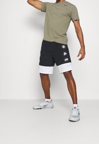 Camiseta verde oliva de manga corta, combinada con pantalones cortos negros con dobladillo blanco y estampado bordado. Zapatillas deportivas grises completan el atuendo.