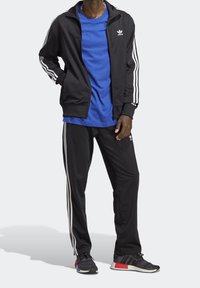 Man i svart Adidas träningsdräkt med vita ränder, en blå t-shirt och svarta sneakers med röda och blå accenter, står med händerna i fickorna.