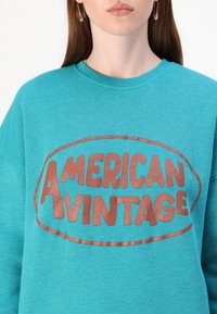 Sweatshirt em teal com um design redondo e o texto "AMERICAN VINTAGE" em letras ousadas de cor cobre. Gola canelada, textura suave.