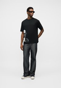 Άνδρας που φοράει μαύρο T-shirt με σχέδιο, σκούρο τζιν, μαύρα γυαλιά ηλίου και μαύρα αθλητικά παπούτσια με λευκή σόλα, στέκεται με το ένα χέρι πίσω από την πλάτη.