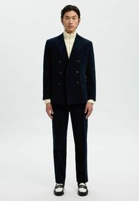 Marineblauer Cordanzug mit doppelter Knopfreihe und einem hochgeschlossenen weißen Pullover, kombiniert mit schwarzen Schuhen mit einem weißen Akzent. Schlichte, maßgeschneiderte Silhouette.