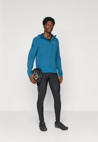 Tyrkysová bunda na zips s kapucňou, čierne priliehavé legíny, čierne cyklistické topánky a čierna prilba, ktorú drží v jednej ruke. Minimalistický dizajn.