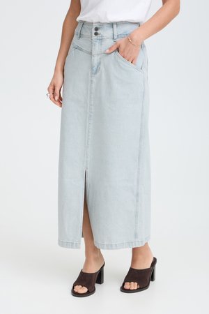 Kvinde iført en lyseblå høj talje denim midi nederdel med frontslids, hvid top, sorte hælede åbne mules og sølvringe på hånden.