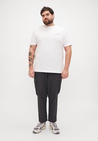 Una camiseta de algodón blanca presenta un pequeño logo, combinada con pantalones cargo oscuros con bolsillos laterales, y zapatillas gris con un diseño estampado.