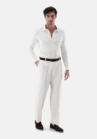 Cremfarbener, gerippter Strickshirt mit einem kragenförmigen Ausschnitt, kombiniert mit weißen, taillierten Hosen und dunkelbraunen Wildleder-Slippern.