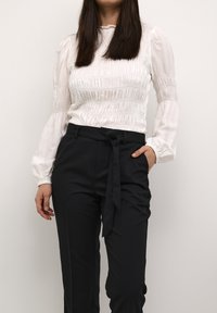 Vit smockblus med långa, insydda ärmar, tillsammans med högmidjade svarta byxor med en knytbältesdetalj.