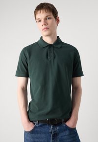 Mørkegrønn polo skjorte laget av myk bomull med klassisk krage, to-knapp placket og korte ermer, sammen med blå denimjeans.